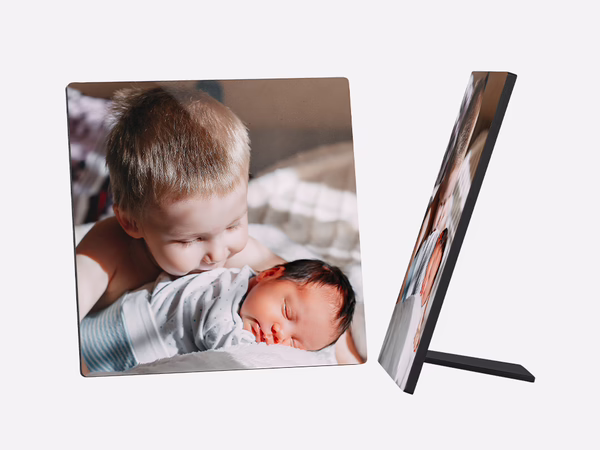 Fotoaufsteller zeigt ein süßes Bild von zwei kleinen Kindern. Ein älteres Kind mit blonden Haaren beugt sich liebevoll über ein neugeborenes Baby, das friedlich schläft. 