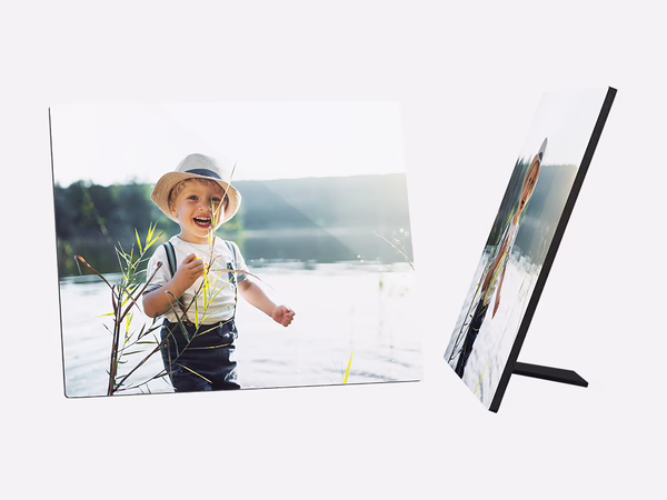 Ein quadratischer Fotoaufsteller zeigt ein fröhliches kleines Kind, das am Ufer eines Sees oder Flusses steht. Das Kind hat blonde Haare und trägt einen hellen Hut, ein weißes T-Shirt mit einem bunten Aufdruck und dunkle Latzhosen. 