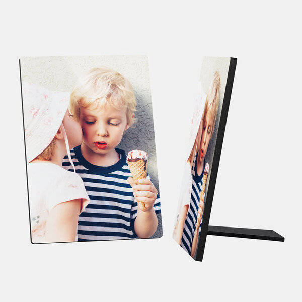 Ein Fotoaufsteller zeigt ein süßes Bild von zwei kleinen Kindern. Ein blondhaariger Junge mit einem blau-weiß gestreiften Shirt hält ein Eis in der Hand und schaut darauf. Ein anderes Kind mit einem hellen Sonnenhut beugt sich zu ihm und scheint ihm einen Kuss auf die Wange zu geben. Der Hintergrund ist hell und schlicht. Rechts daneben ist ein weiterer Fotoaufsteller mit dem gleichen Motiv zu sehen, der auf seiner Stütze steht.